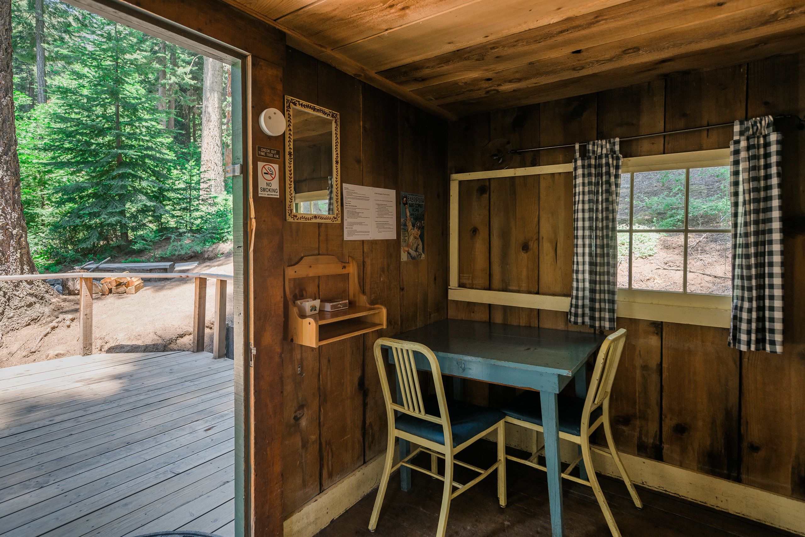 Una cabaña con interior de madera, una mesa con dos sillas, un espejo y una ventana con cortinas de cuadros.