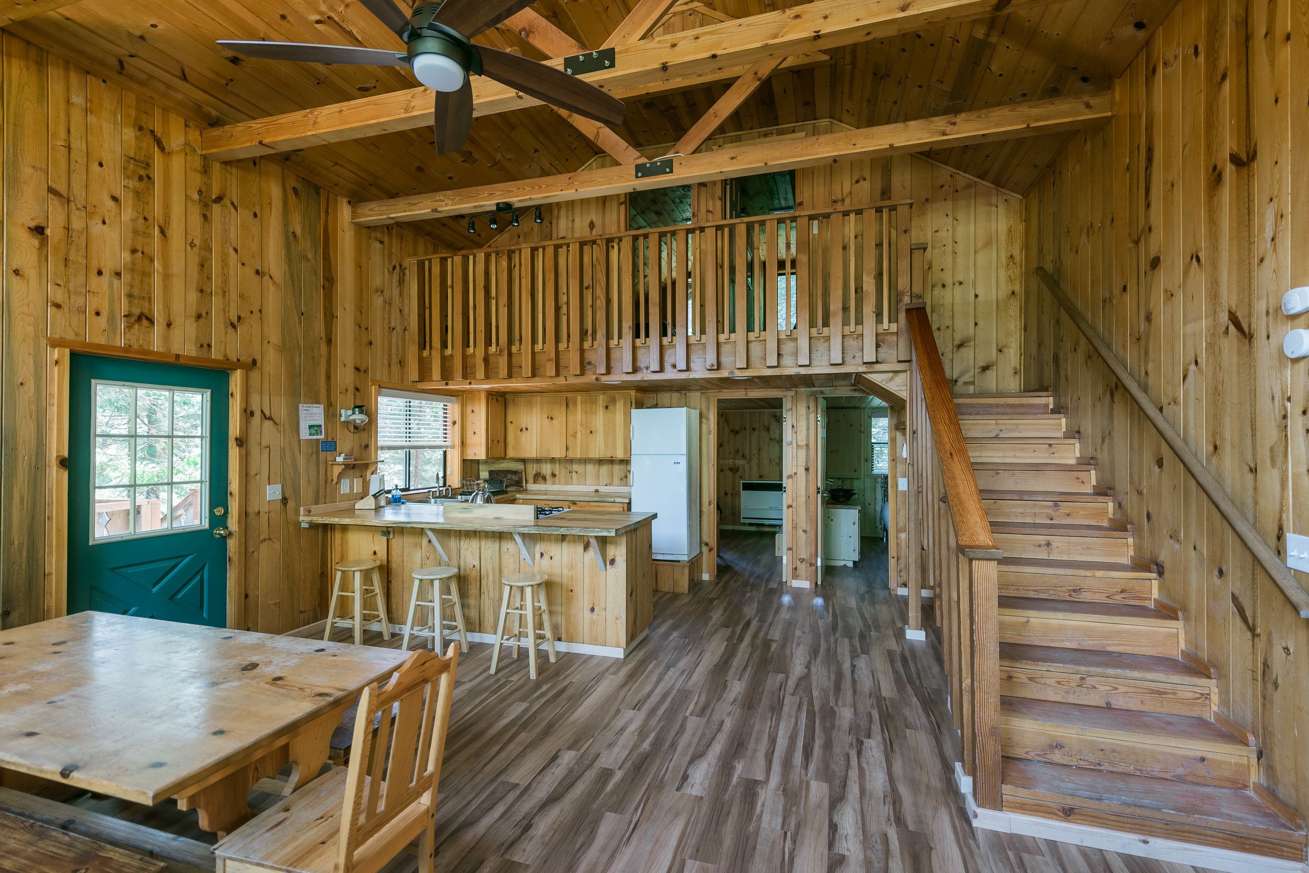 A wooden cabin interior with a kitchen, dining area, and a staircase leading to a loft.
