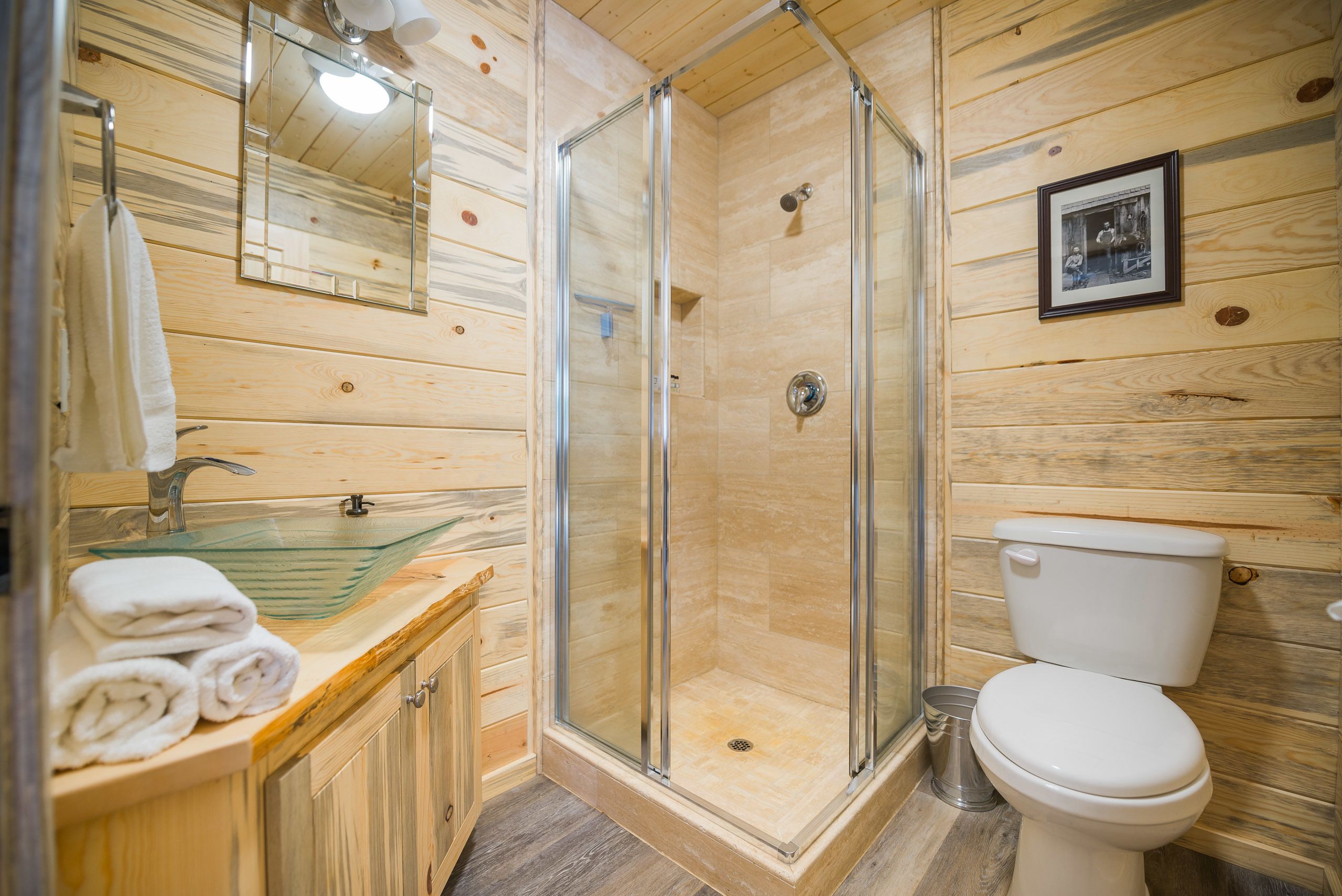 A wooden cabin bathroom with a glass shower enclosure, a sink with a glass basin, and a toilet.