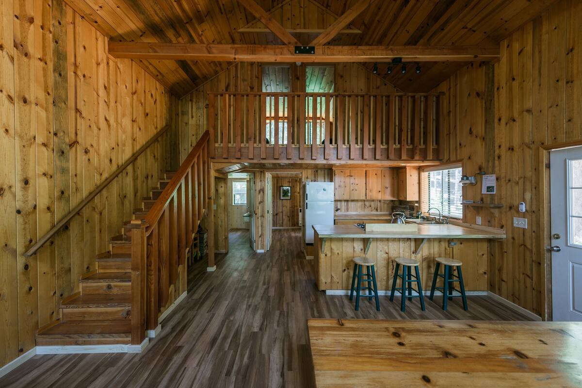 A wooden cabin interior with a central staircase, a kitchen area with a bar, and a dining space.