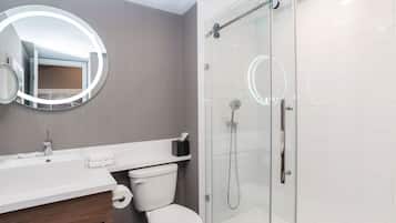 Un baño moderno con ducha de vidrio, un espejo redondo con luz y un lavamanos blanco.