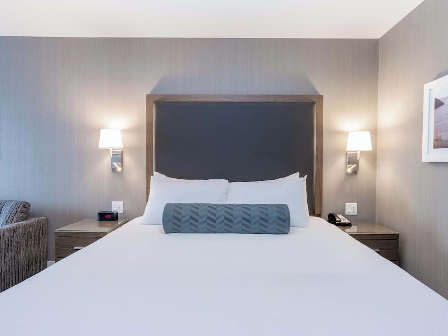 Habitación de hotel con una cama grande, dos mesitas de noche, una silla y una mesita auxiliar con lámpara.