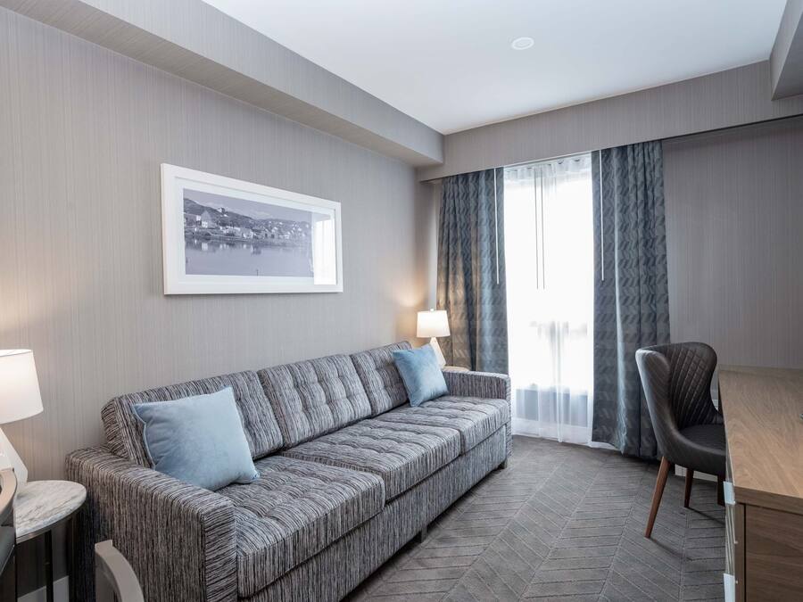 A hotel room with a sofa, a chair, a desk, a lamp, a picture on the wall, and a window with curtains.