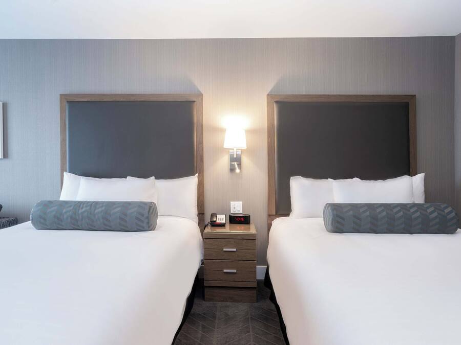 A hotel room with two beds, a nightstand with a lamp, and a small table with a phone and clock.