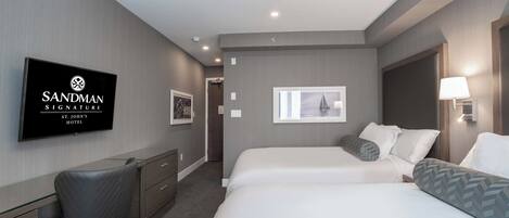 A hotel room with a bed, a desk, a chair, a TV displaying the hotel name, and a window with a sailboat view.