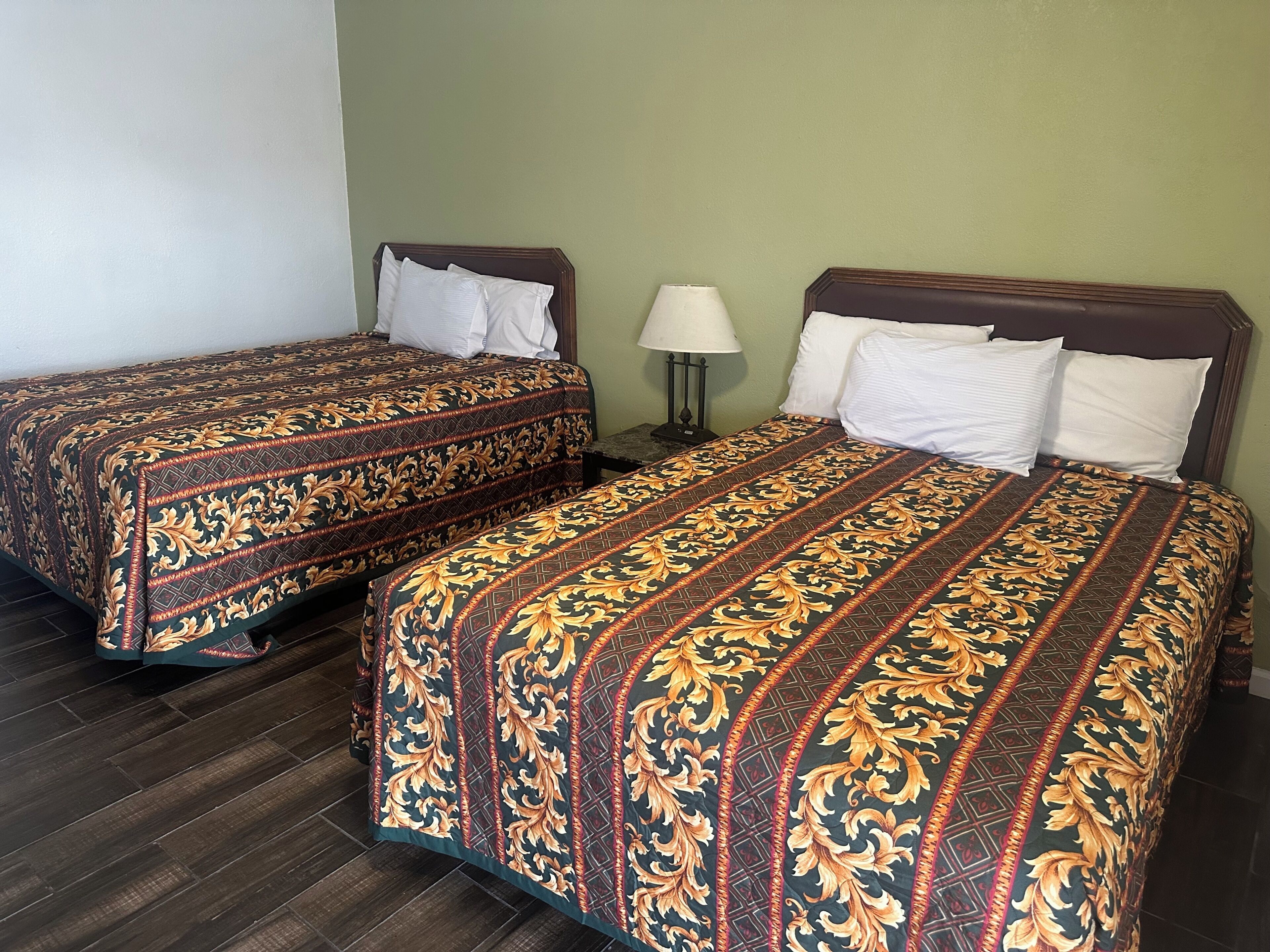 Dos camas individuales con ropa de cama estampada, una mesita de noche con lámpara y una pared verde.