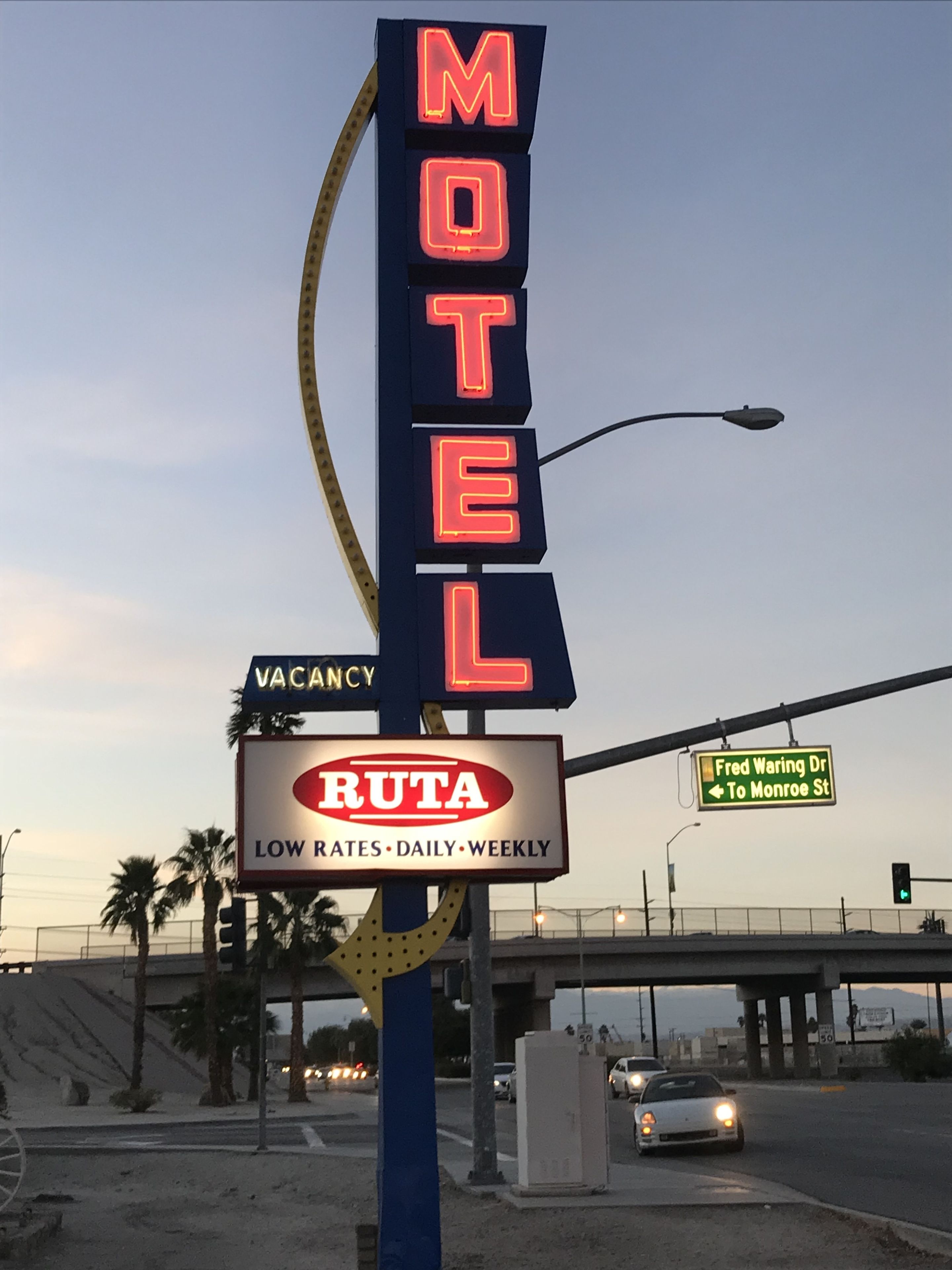 Een motelbord met neonverlichting waarop 'MOTEL' staat en een bord eronder met de tekst 'RUTA SCHERP TARIEVEN DAGELIJKS WEEKLIJKS'.