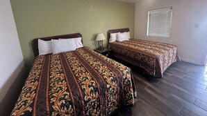 Dos camas con ropa de cama estampada, una mesita de noche con lámpara, una ventana con persianas y una luz en el techo.