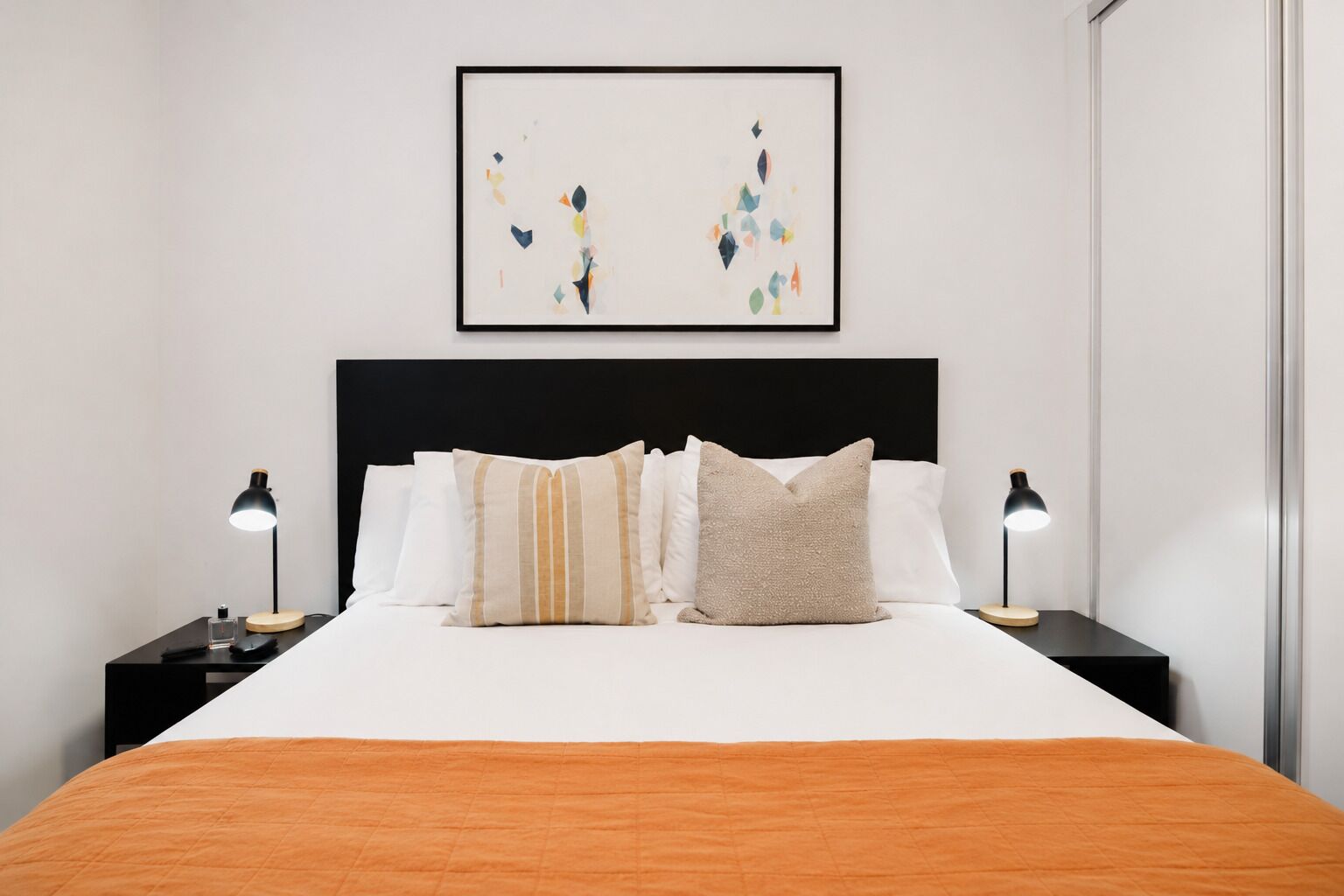 A modern bedroom with a large bed, two bedside tables with lamps, a black headboard, and a framed abstract artwork above the bed.