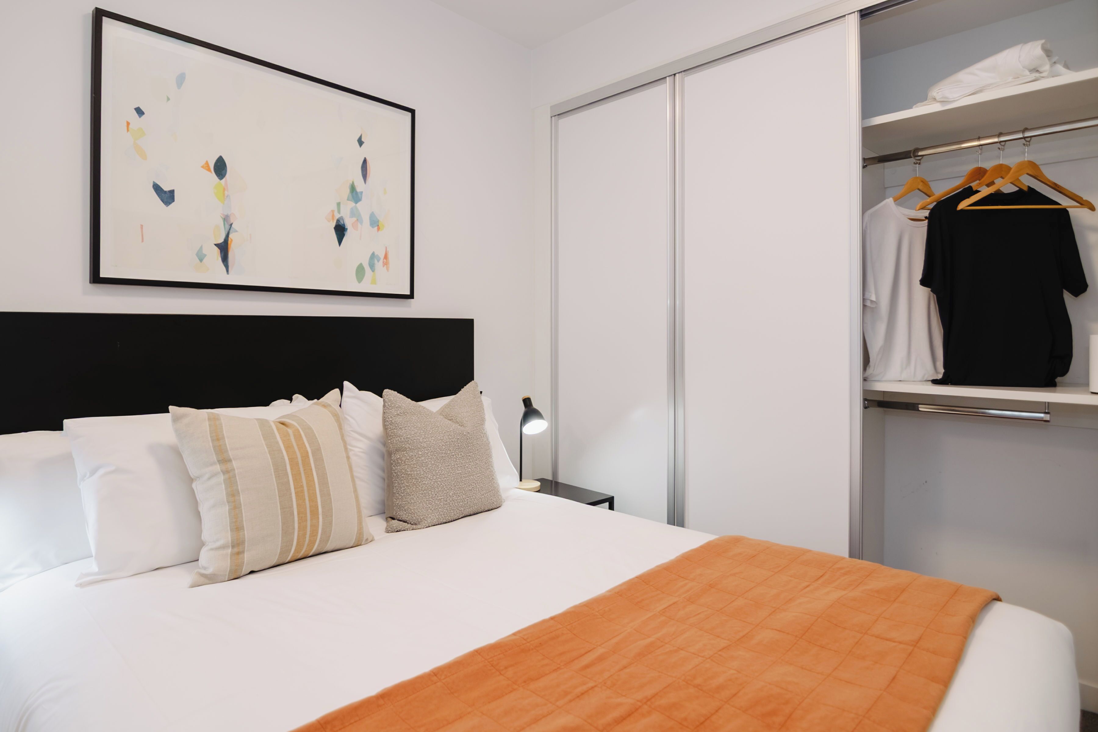 A hotel room with a bed, a bedside table, a lamp, a wardrobe, and a clothing rack.