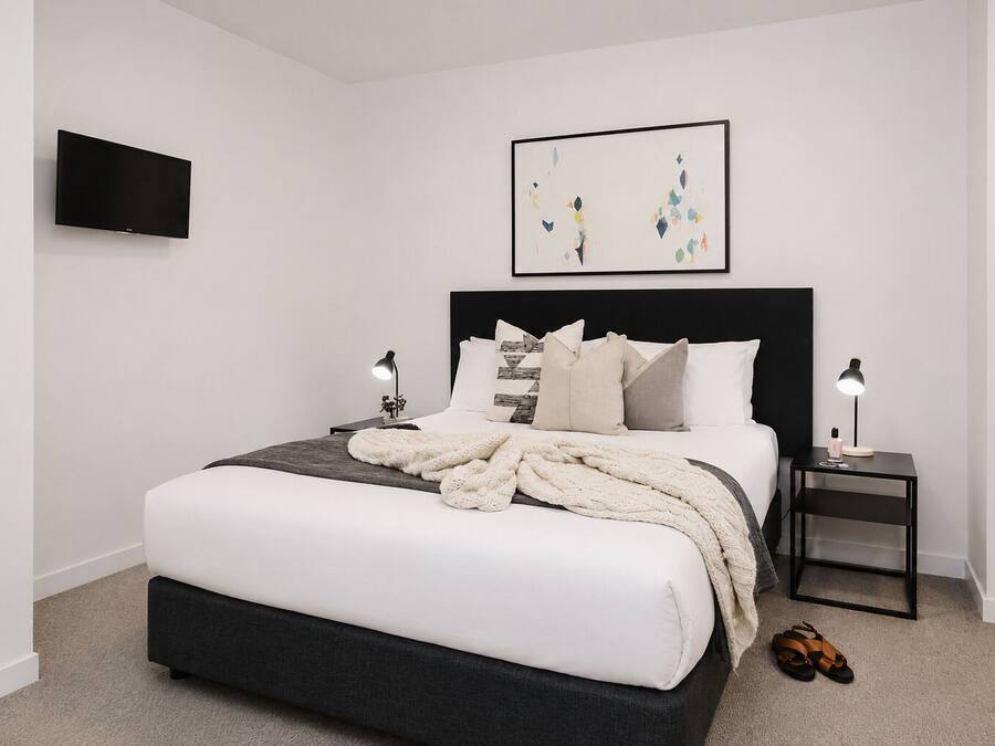 A modern bedroom with a large bed, a flat-screen TV, a framed abstract painting, two bedside tables with lamps, and a pair of shoes on the floor.