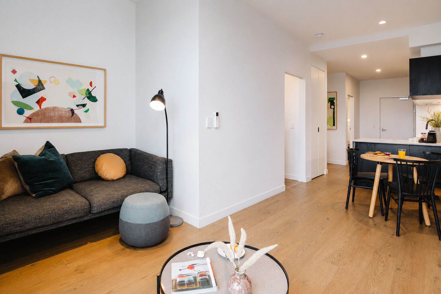 Una sala de estar moderna con sofá, reposapiés y una mesa de centro con un florero y un libro. Se observa un comedor con mesa y sillas, y una cocina al fondo.