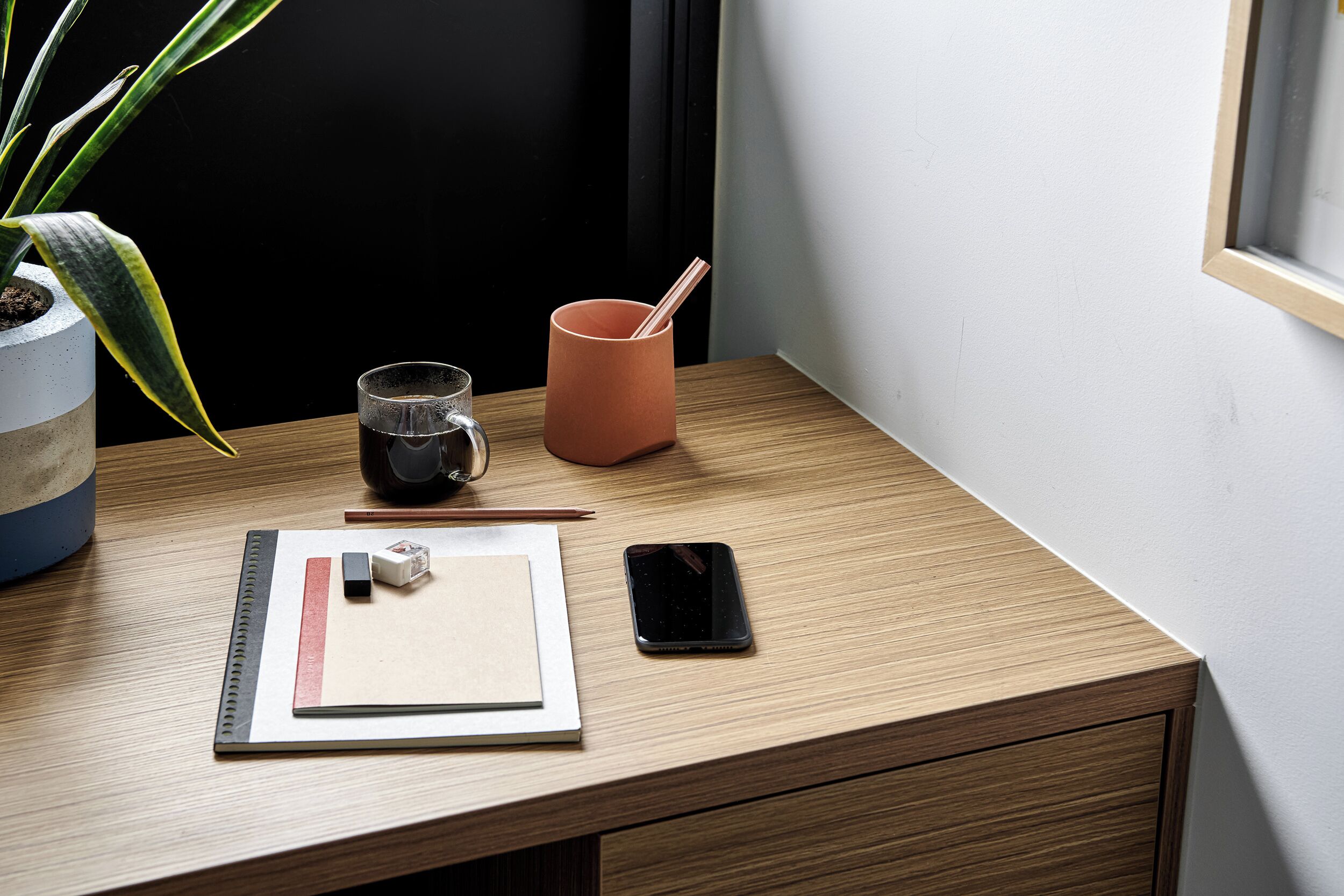 Et træskrivebord med en smartphone, et glas vand, en blyant, en lineal og en lille plante.