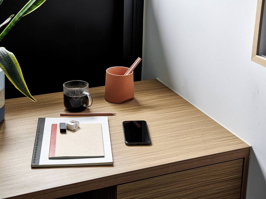 Et træskrivebord med en smartphone, et glas vand, en blyant, en lineal og en lille plante.