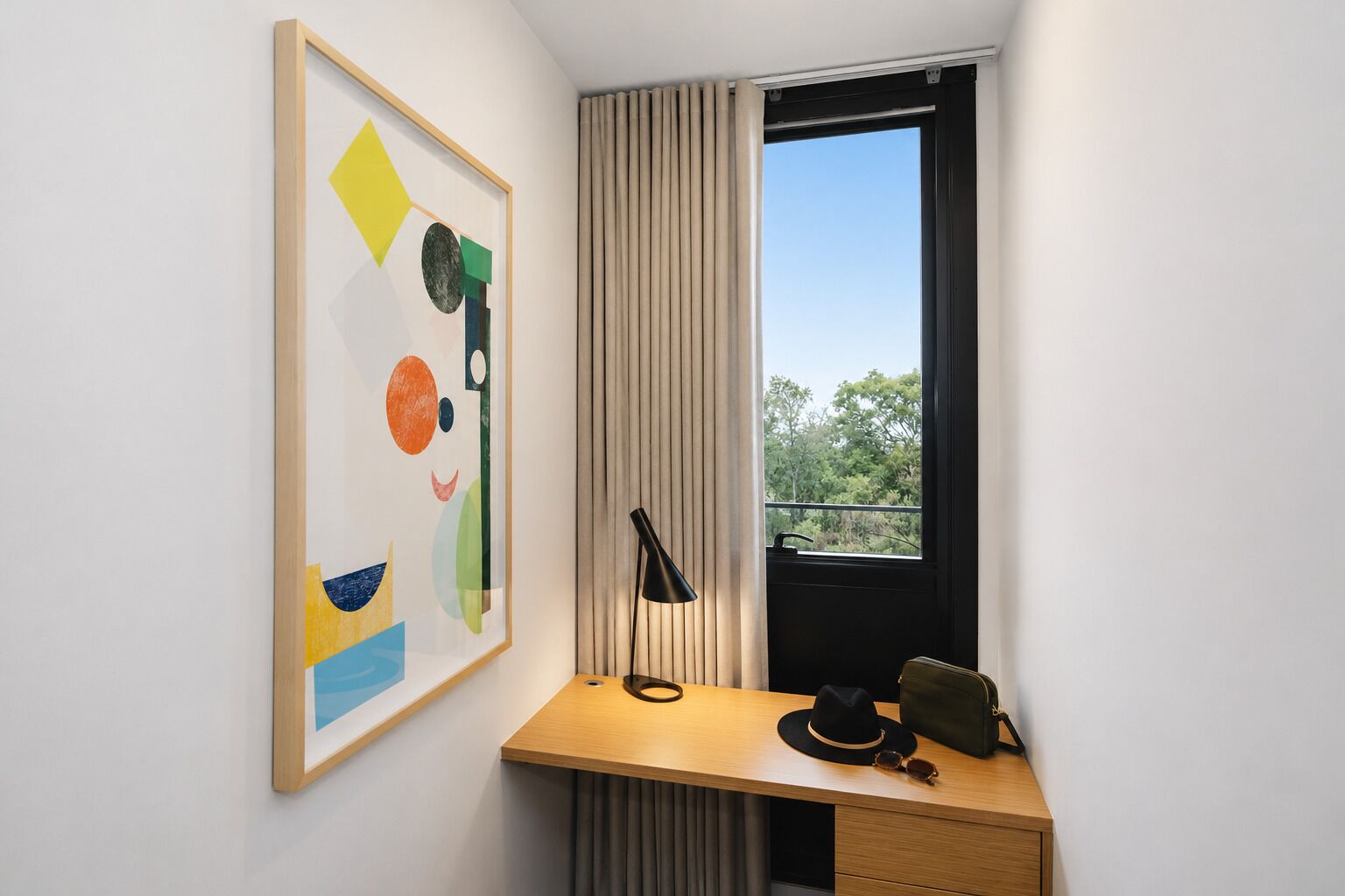 A room with a desk, a framed abstract painting, a window with curtains, and a view of trees.