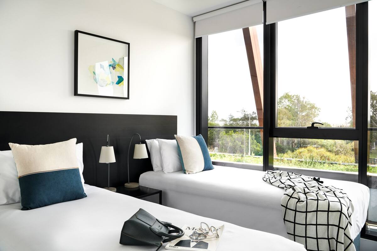 Habitación de hotel con dos camas, un cuadro abstracto enmarcado, una ventana con vistas a zonas verdes y una mesita de noche con lámpara.