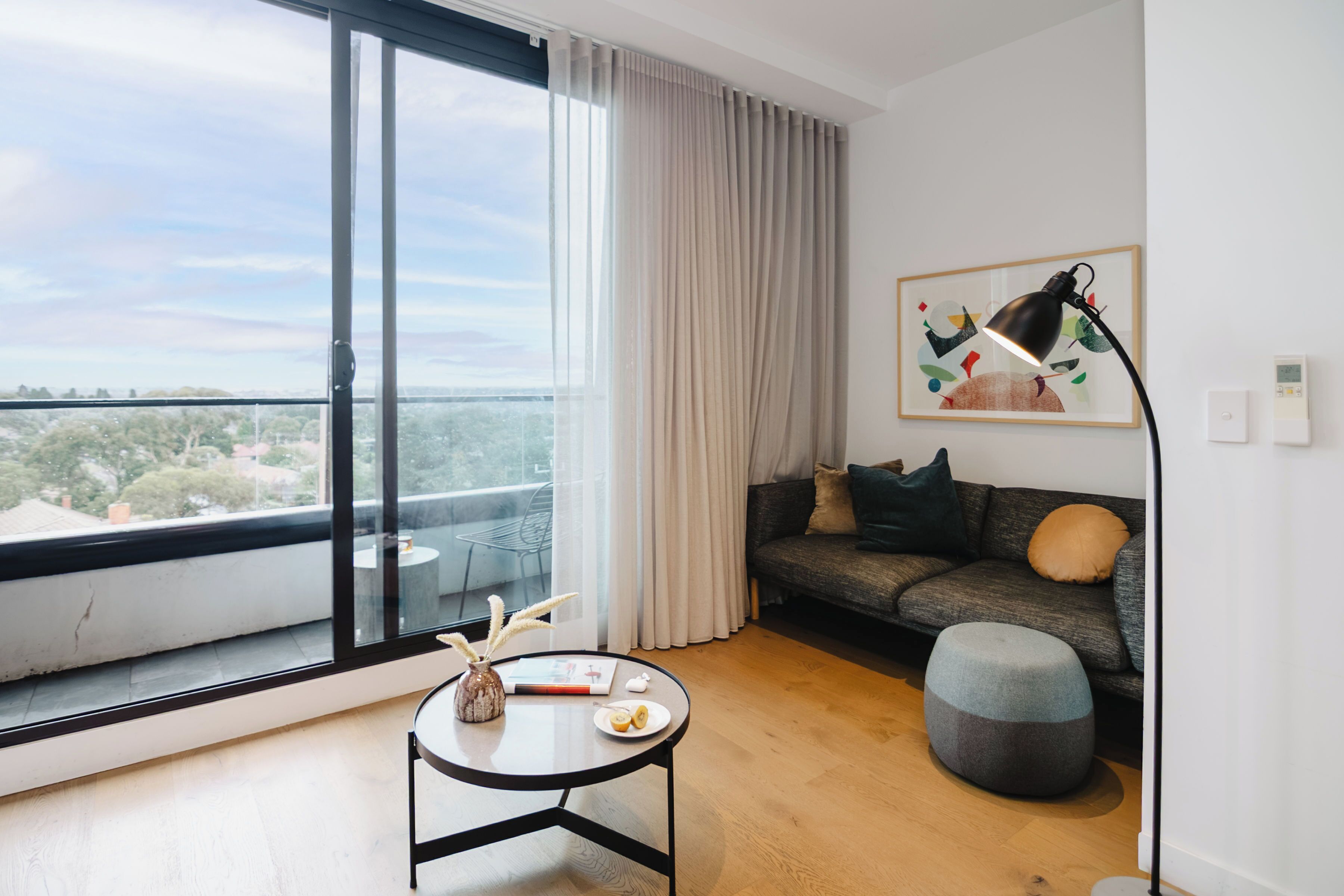 A modern living room with a large window, a sofa, a round table, and a lamp.