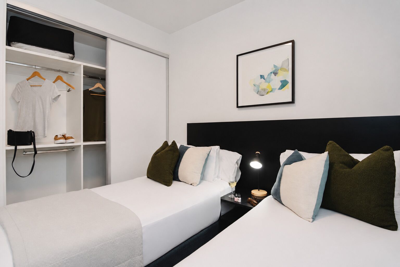 A hotel room with two beds, a wardrobe, a bedside table with a lamp, and a framed abstract artwork on the wall.