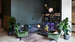 Una sala de estar moderna con una pared azul oscuro, sillones verdes, un sofá gris y una mesa de centro. Se observa una estantería con diversos objetos y una planta grande en maceta.