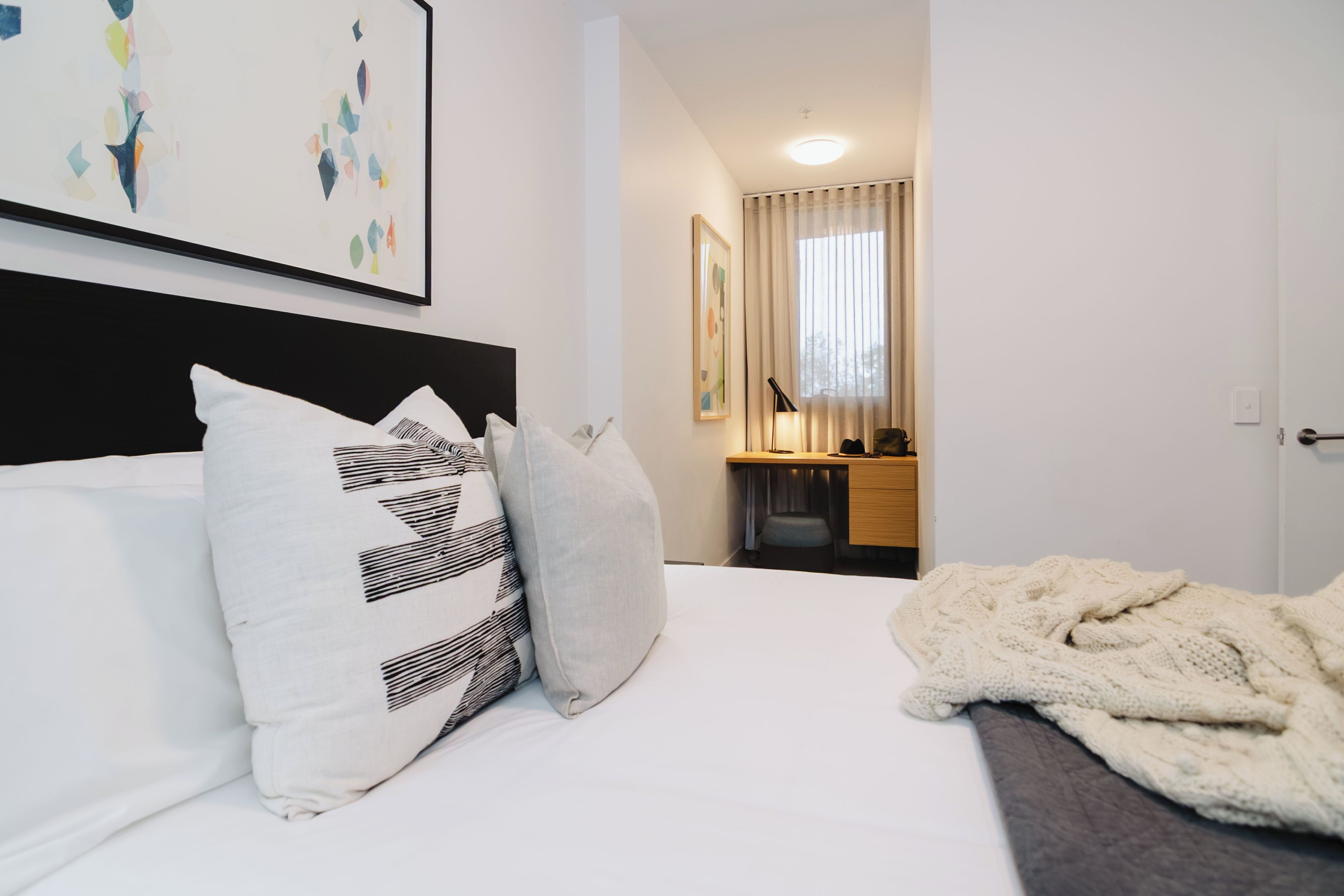 A hotel room with a bed, pillows, a blanket, a desk with a chair, a mirror, and artwork on the wall.