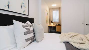 A hotel room with a bed, pillows, a blanket, a desk with a chair, a mirror, and artwork on the wall.
