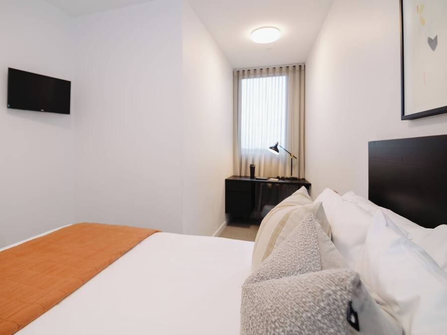 Et hotelværelse med seng, skrivebord, stol, fjernsyn og et vindue med gardiner.