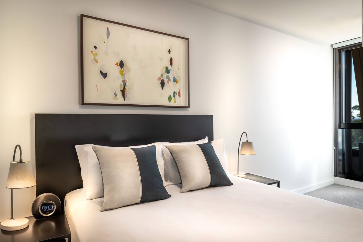 Habitación de hotel moderna con una cama grande, dos mesitas de noche, un reloj de pared y un cuadro abstracto enmarcado.