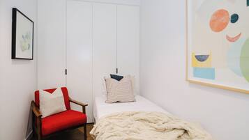 A bedroom with a single bed, a red chair, a white wardrobe, and a large abstract painting.