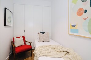 Un dormitorio con una cama individual, una silla roja, un armario blanco y un cuadro abstracto grande.