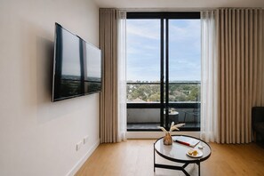 A modern living room with a flat-screen TV mounted on the wall, a round table with a vase and a plate, and a balcony with a view of the city. - Punthill Ivanhoe (Ivanhoe)