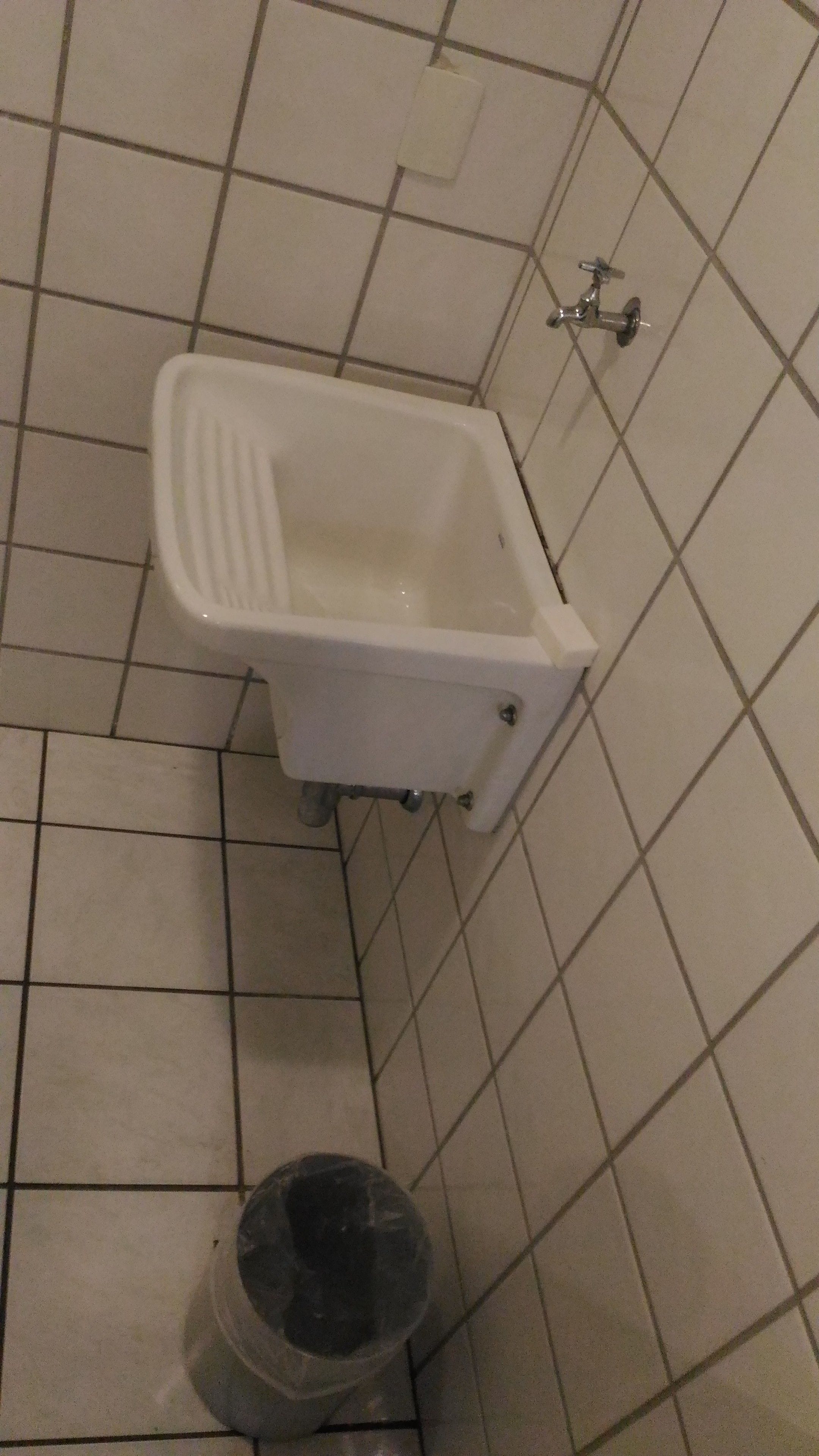 A small, white, square-shaped urinal with a metal handle, mounted on a tiled wall.