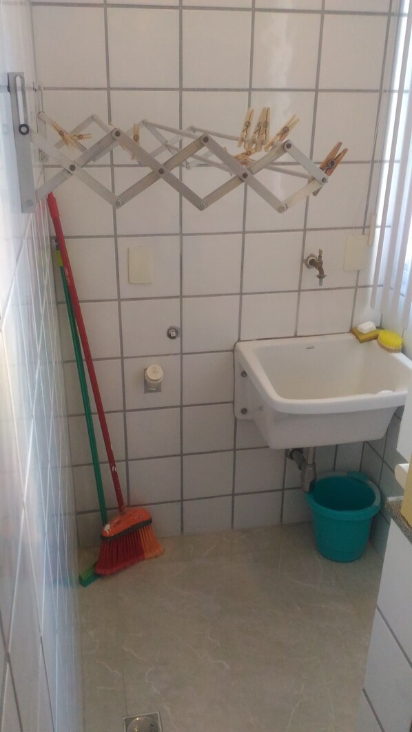 A bathroom with a tiled floor, a sink, a blue bucket, a broom, and a drying rack with clothespins.