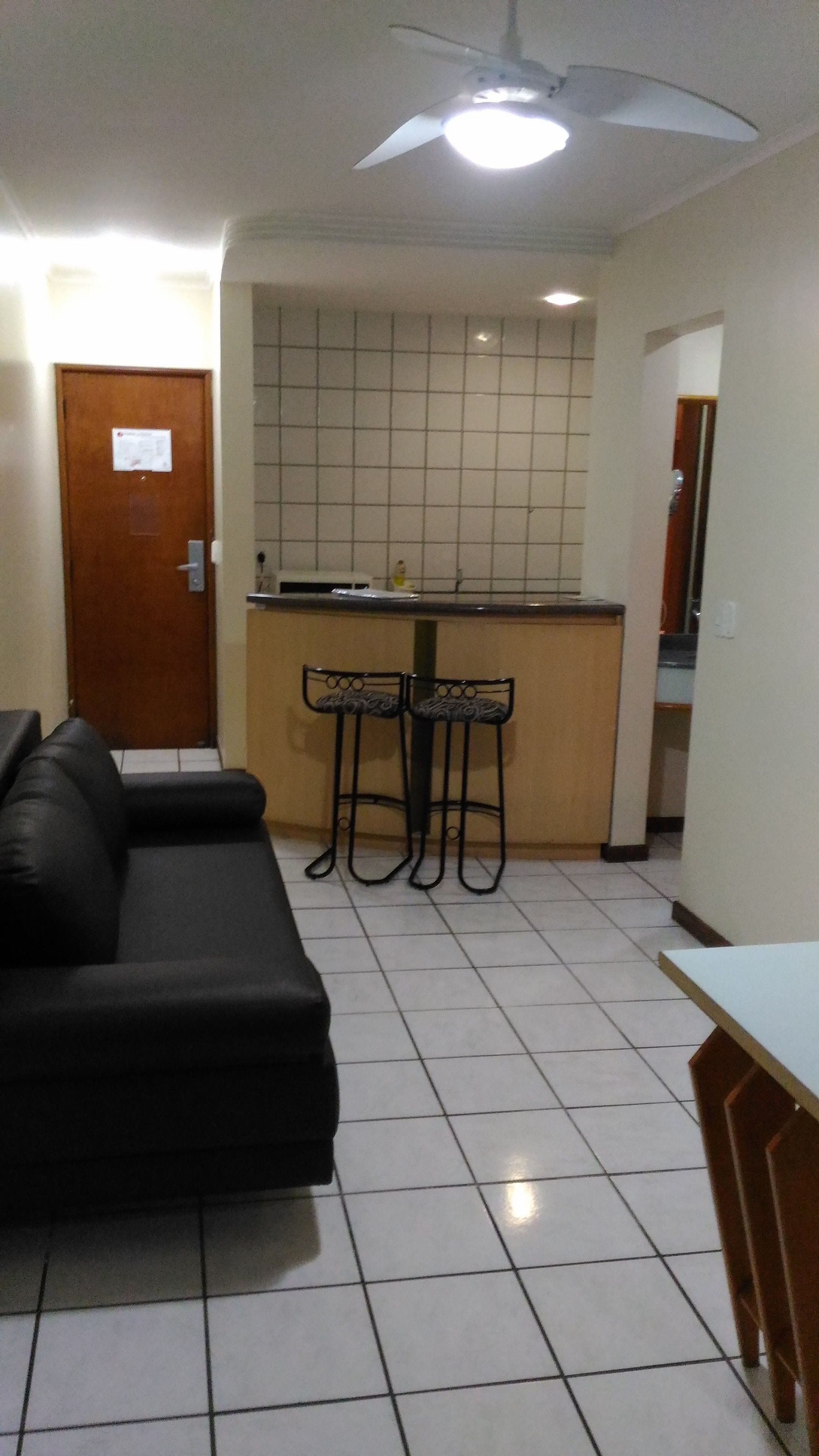 Sala de estar com sofá preto, piso ladrilhado, ventilador de teto e área de cozinha com bancada e banquetas.