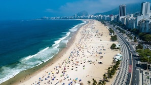 A beach with people and umbrellas, a road alongside, and buildings in the background. - Copacabana Near the Beach (Rio de Janeiro)
