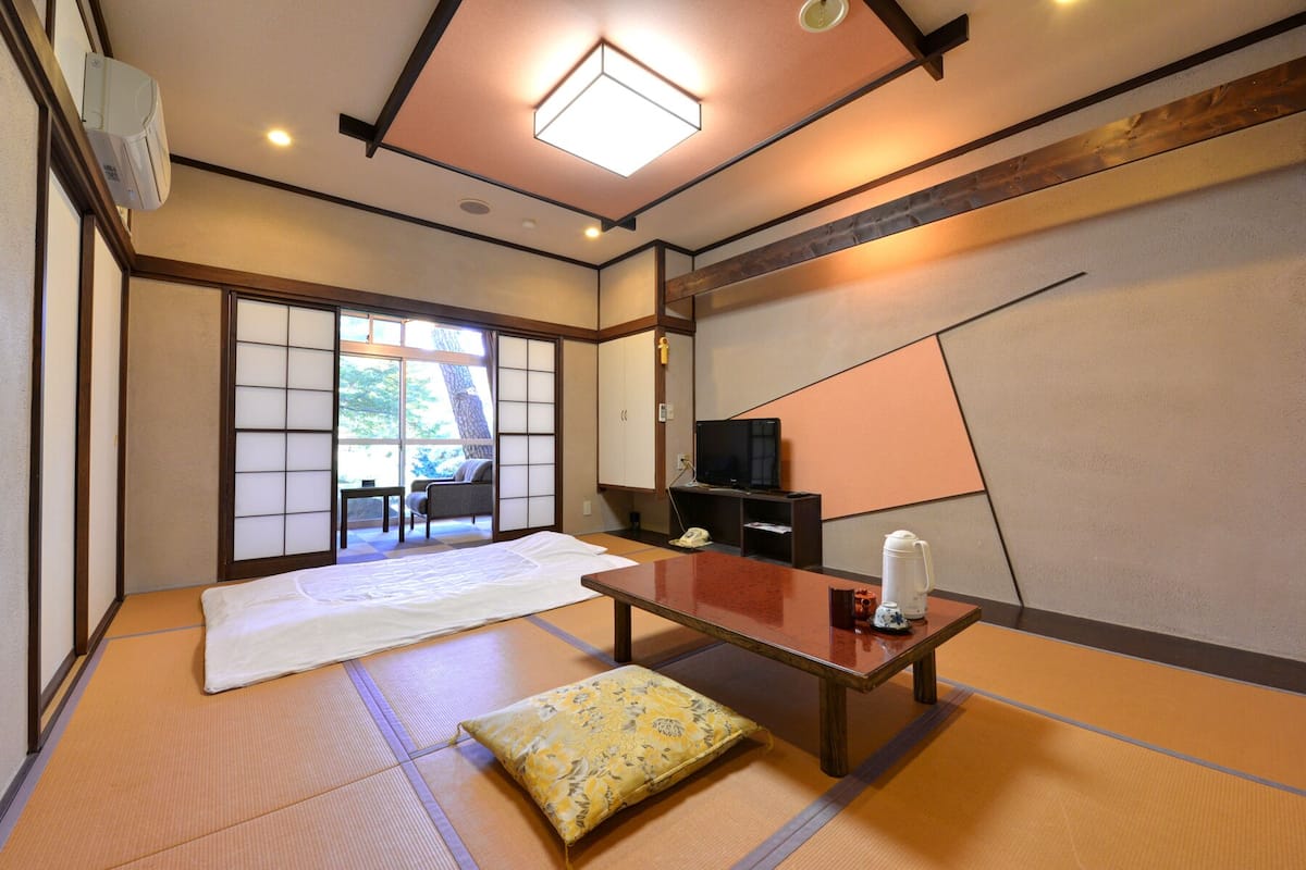 A traditional Japanese room with tatami flooring, a low table, a television, and sliding doors leading to an outdoor area.