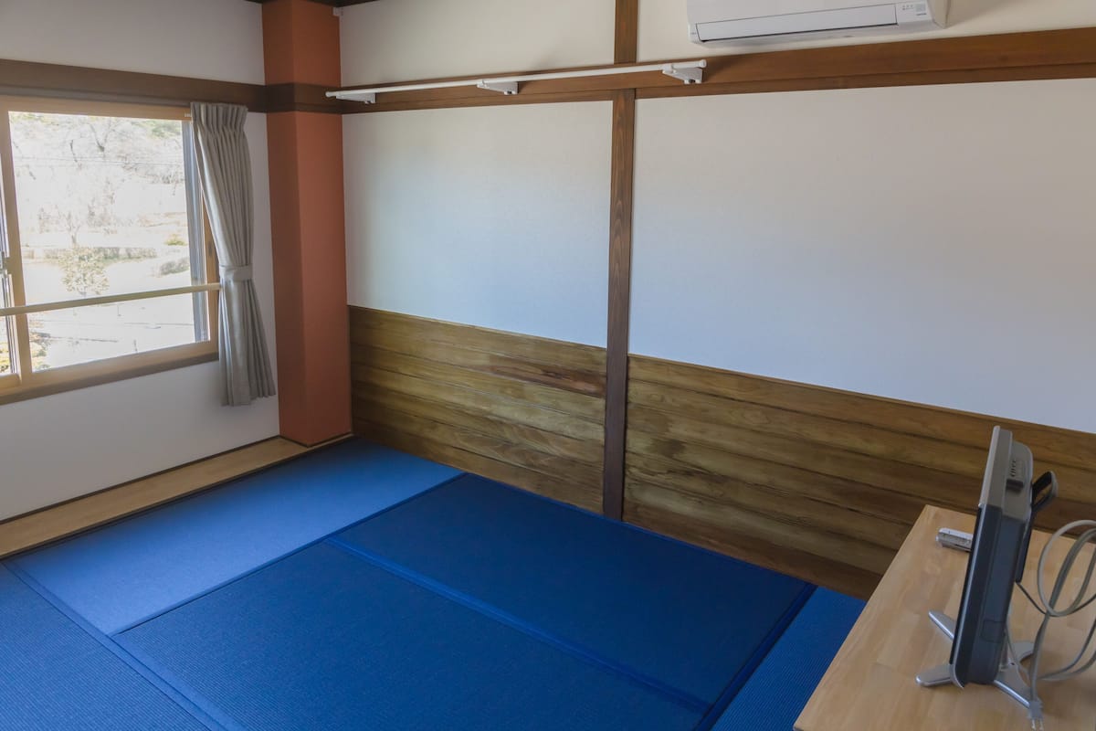 A room with tatami mats, a wooden beam ceiling, a window with curtains, and a desk with a device.