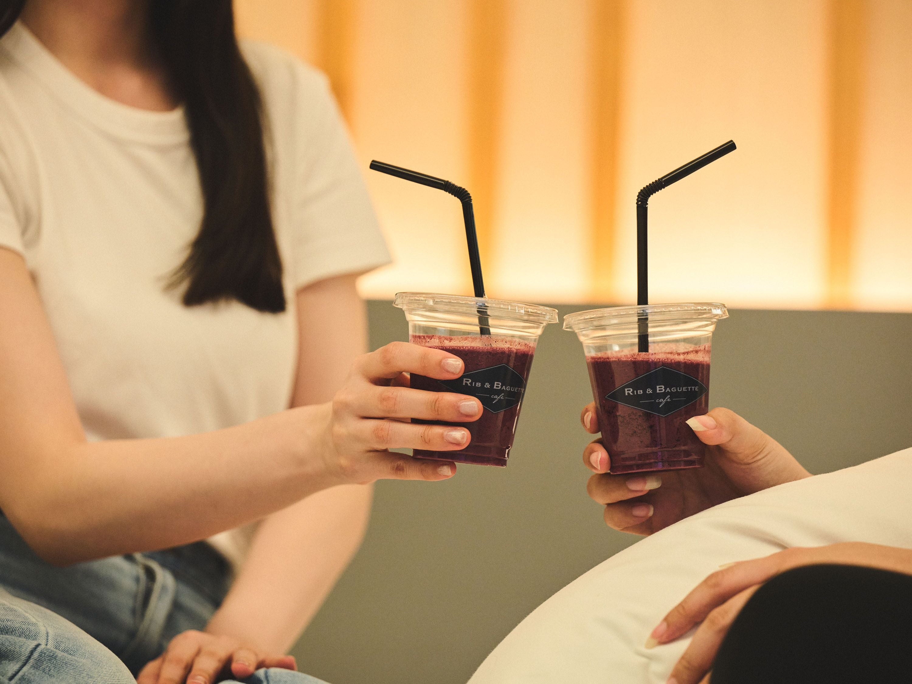 Two people holding drinks with straws, one with a label that reads 'Rib & Baguette cafe'.