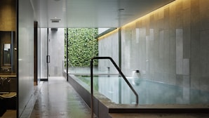 Un área de piscina interior moderna con una pared de vidrio, una escalera de metal y vistas a zonas verdes exteriores.