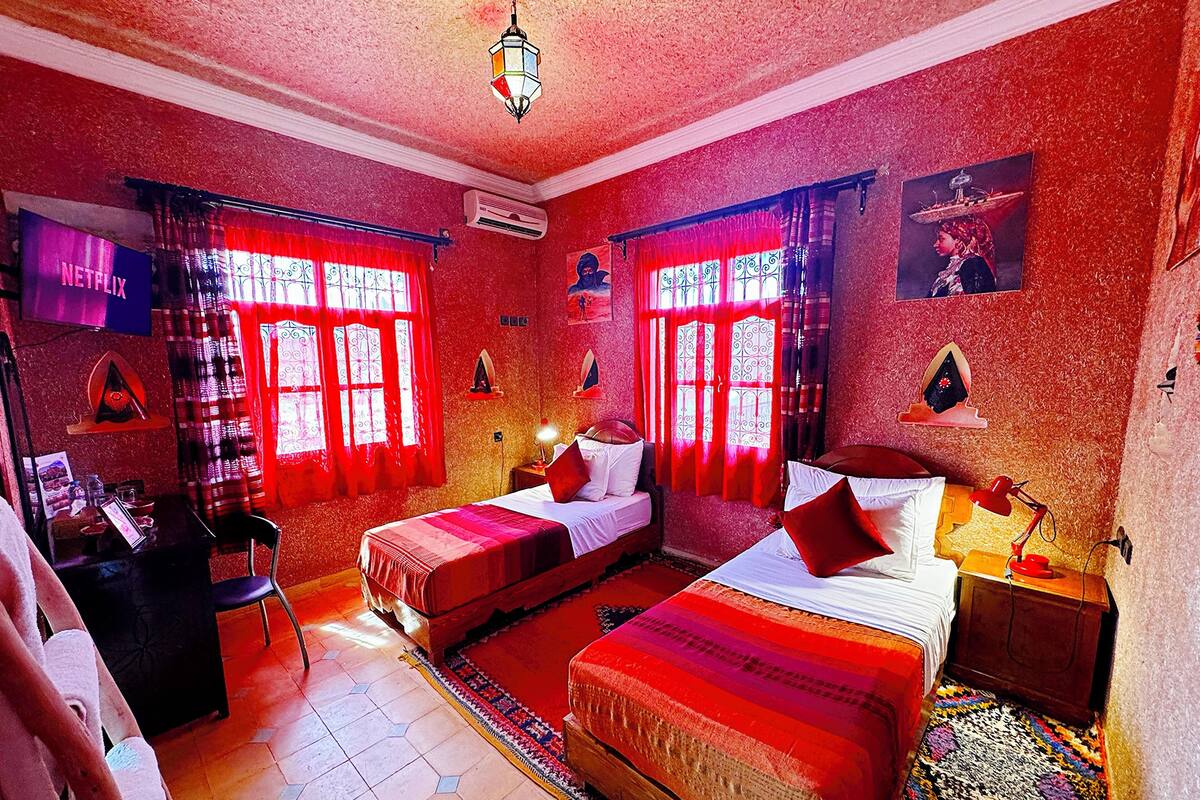Ein Zimmer mit zwei Betten, einem Schreibtisch und einem Stuhl. Es gibt zwei Fenster mit roten Vorhängen.