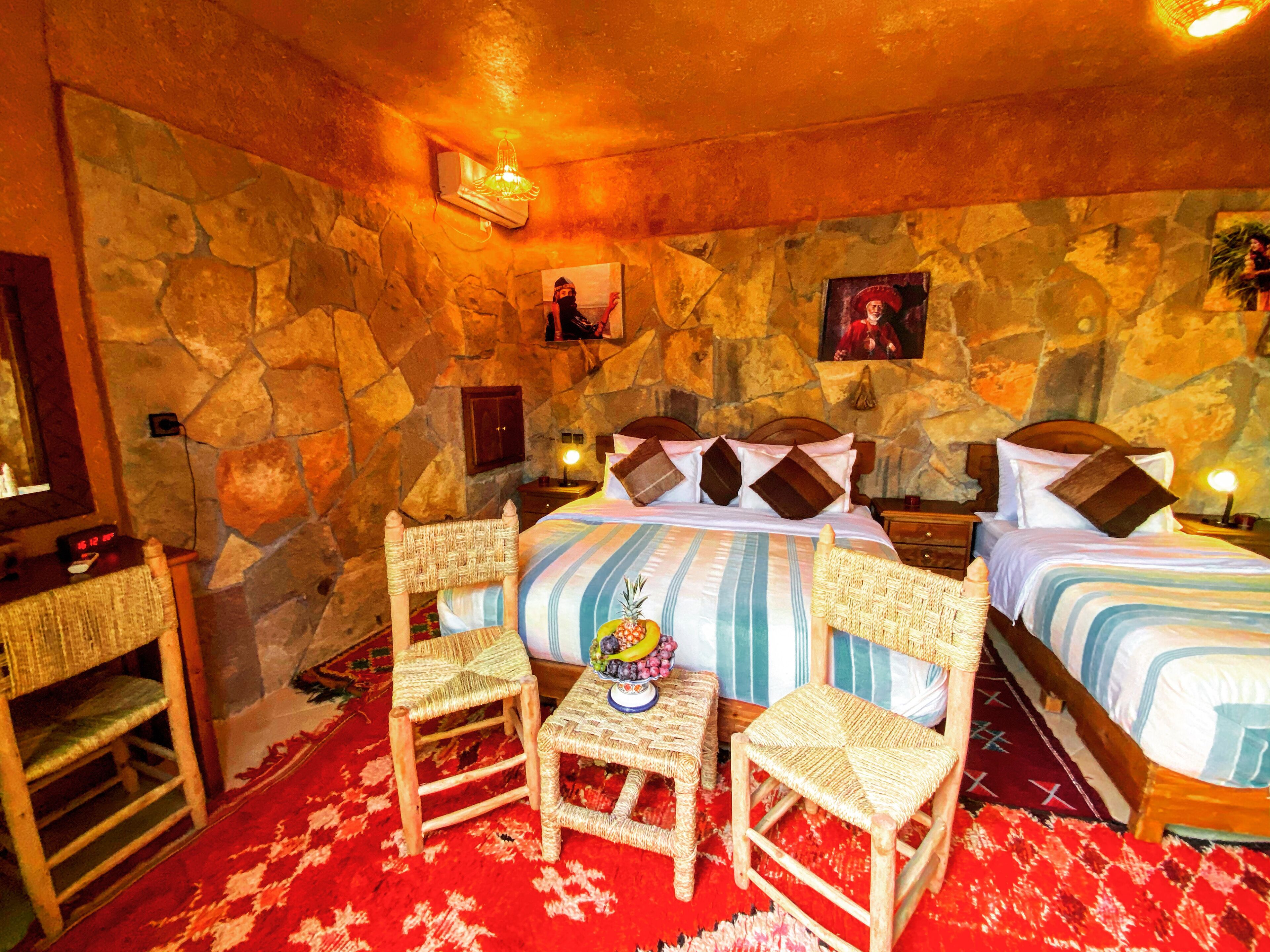 Ein Zimmer mit Steinwand, einem Bett mit gestreifter Bettwäsche, zwei Korbsesseln, einem kleinen Tisch mit Obst und einem Nachttisch mit Lampe.