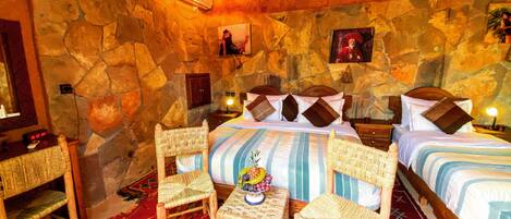 A room with a stone wall, a bed with striped bedding, two wicker chairs, a small table with fruit, and a nightstand with a lamp.