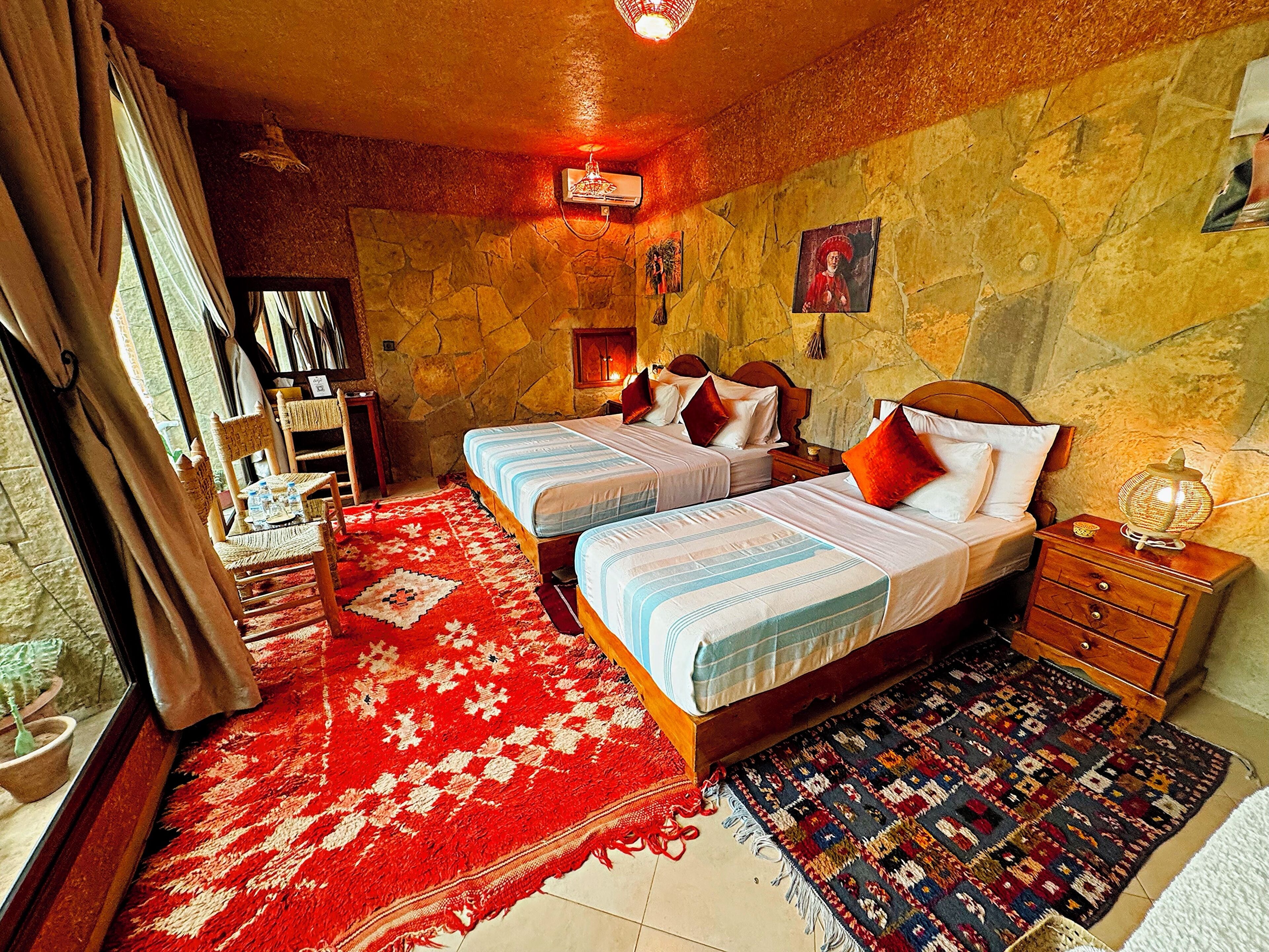 Quarto com duas camas, um tapete estampado em vermelho e azul, uma mesinha de cabeceira de madeira e uma parede de pedra.