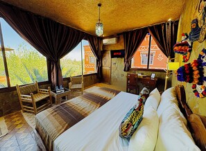A room with a large bed, a desk, and a chair. There is a window with curtains, a hanging light fixture, and a wall with a colorful decoration.