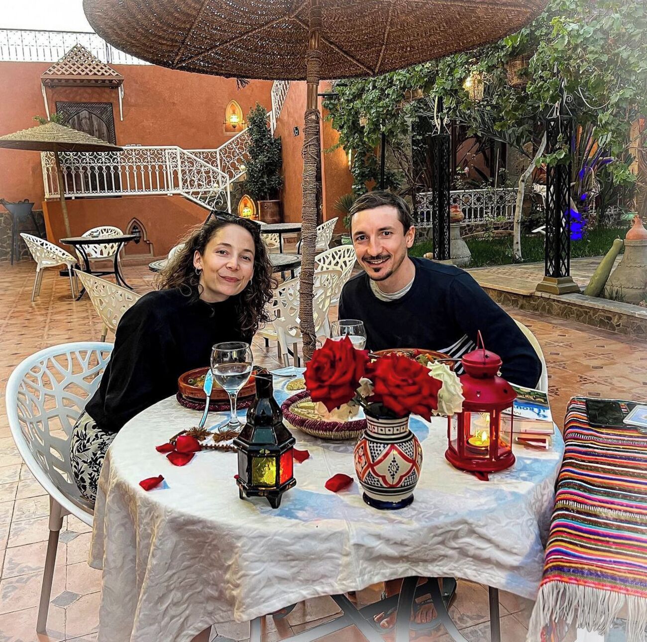 Mesa posta ao ar livre com toalha branca, rosas vermelhas e lanternas.