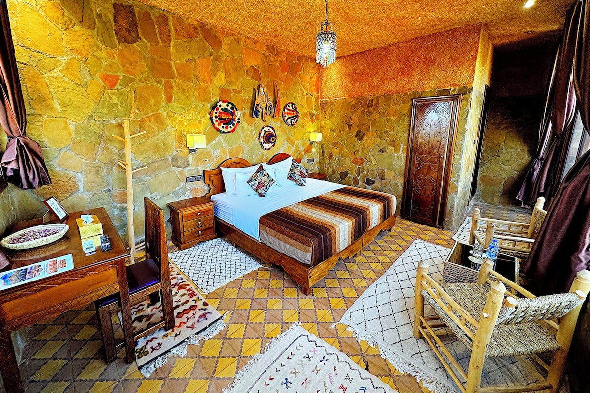 Ein traditionell eingerichtetes Zimmer mit Steinwand, einem Bett mit gemusterten Textilien, einem hölzernen Nachttisch, einem Sessel und einer Pendelleuchte.