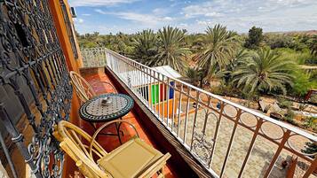 Un balcón con mesa y sillas, baranda decorativa y vista a palmeras y follaje.