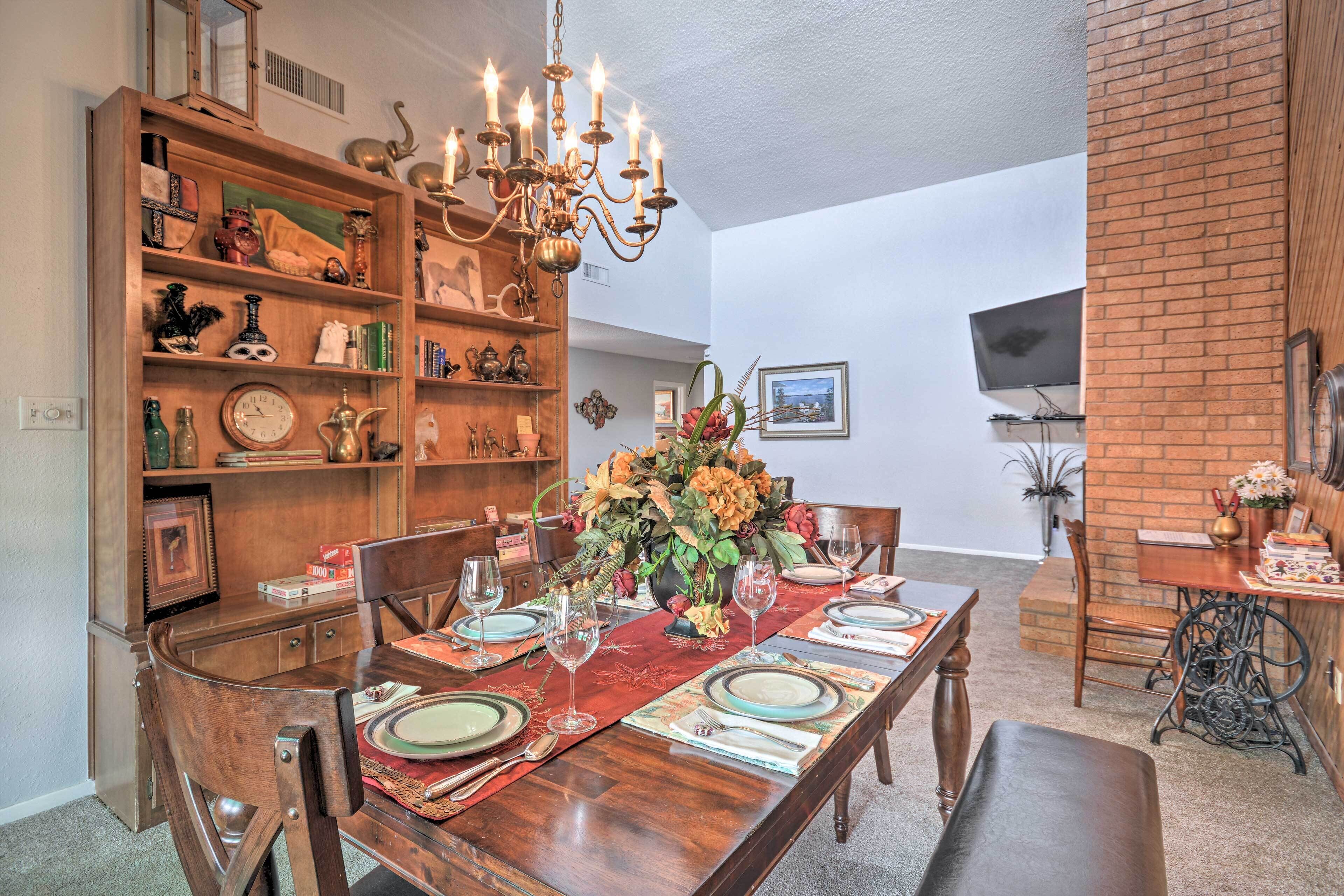 Comedor con una mesa de madera dispuesta para seis personas, rodeada de sillas. Un candelabro cuelga sobre la mesa, y un estante lleno de diversos objetos se encuentra a la izquierda.
