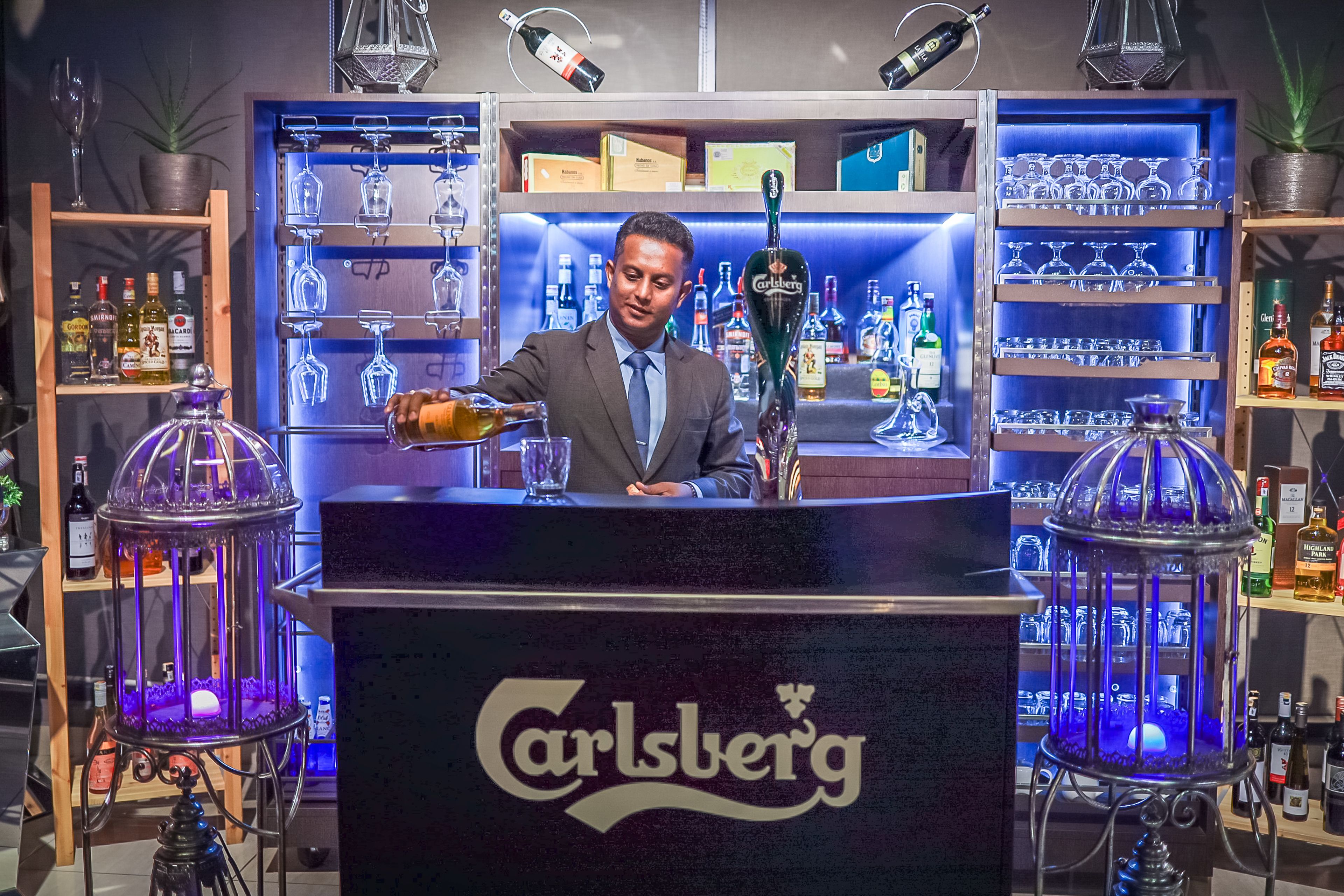 Zona de bar con un barman, estantes llenos de botellas de licor y copas iluminadas.