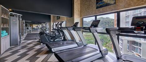 Un gimnasio con cintas de correr, máquinas de pesas y vistas a un edificio a través de amplias ventanas.