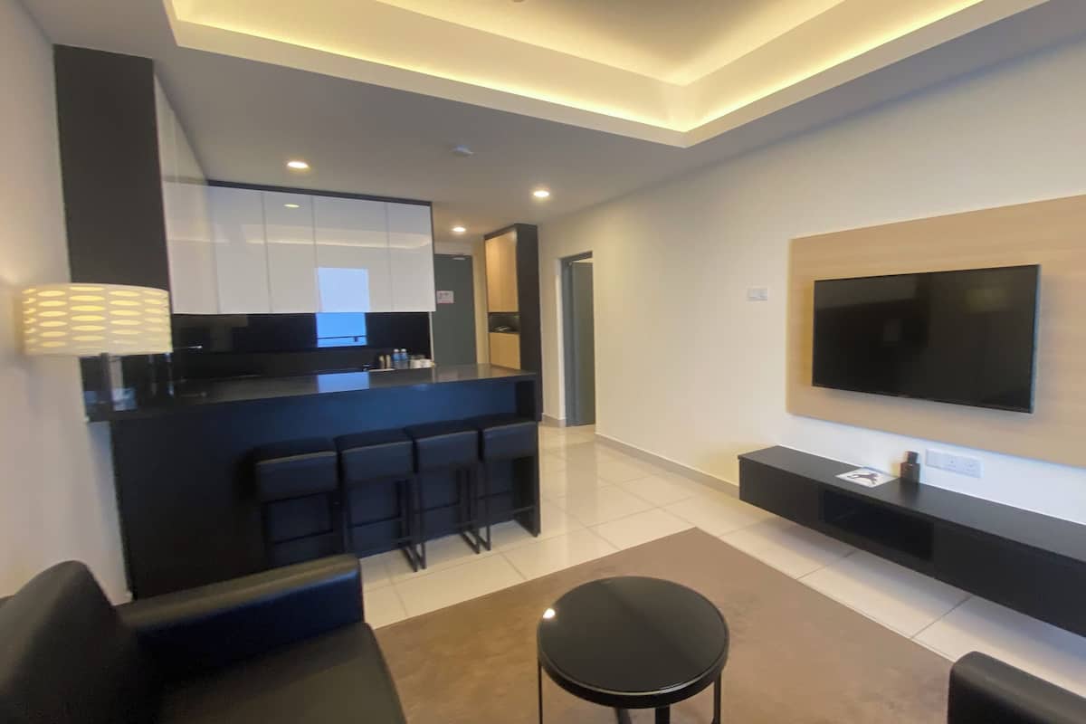 Un salon moderne avec un grand téléviseur à écran plat fixé au mur, un canapé noir, une petite table ronde et un coin cuisine doté d’un plan de travail noir et de tabourets de bar.